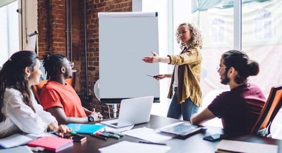 People sitting at a table looking at a flip chart and a person presenting next to it